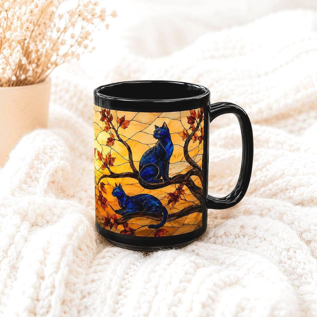A black ceramic mug with a colorful illustration of two blue cats sitting on tree branches with orange leaves, placed on a white knit blanket.