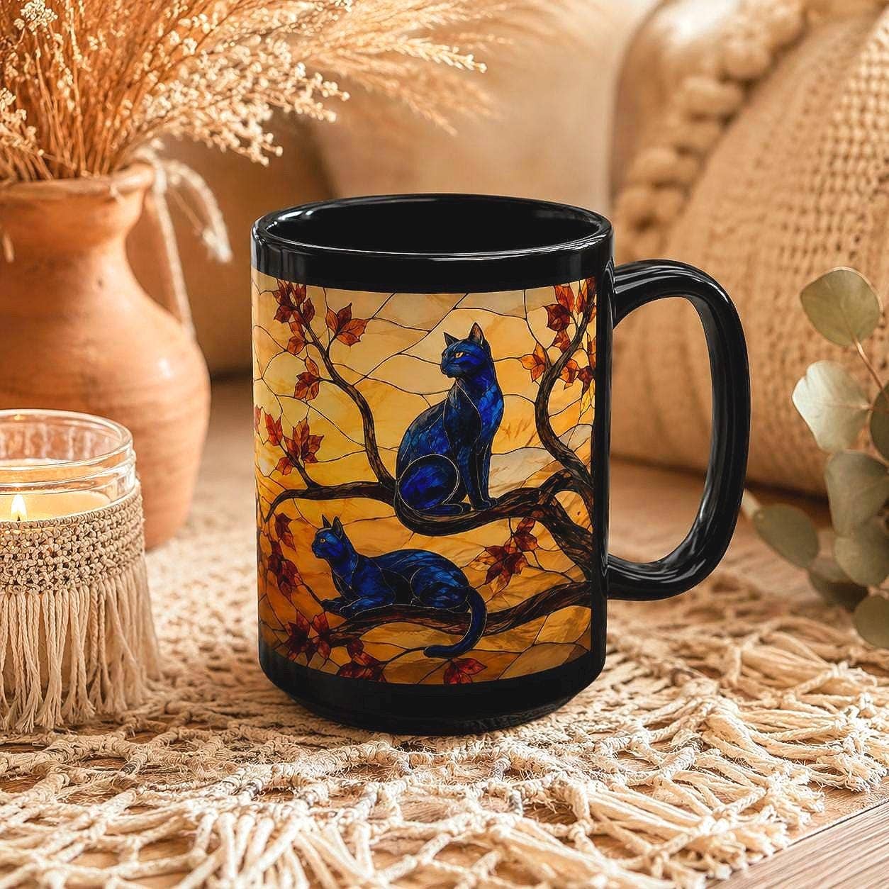 A ceramic mug with a black handle and rim, featuring a stained-glass-style illustration of two blue cats on branches with red leaves against a yellow background. The mug is styled on a woven mat with a candle, dried flowers, and a cozy beige background.