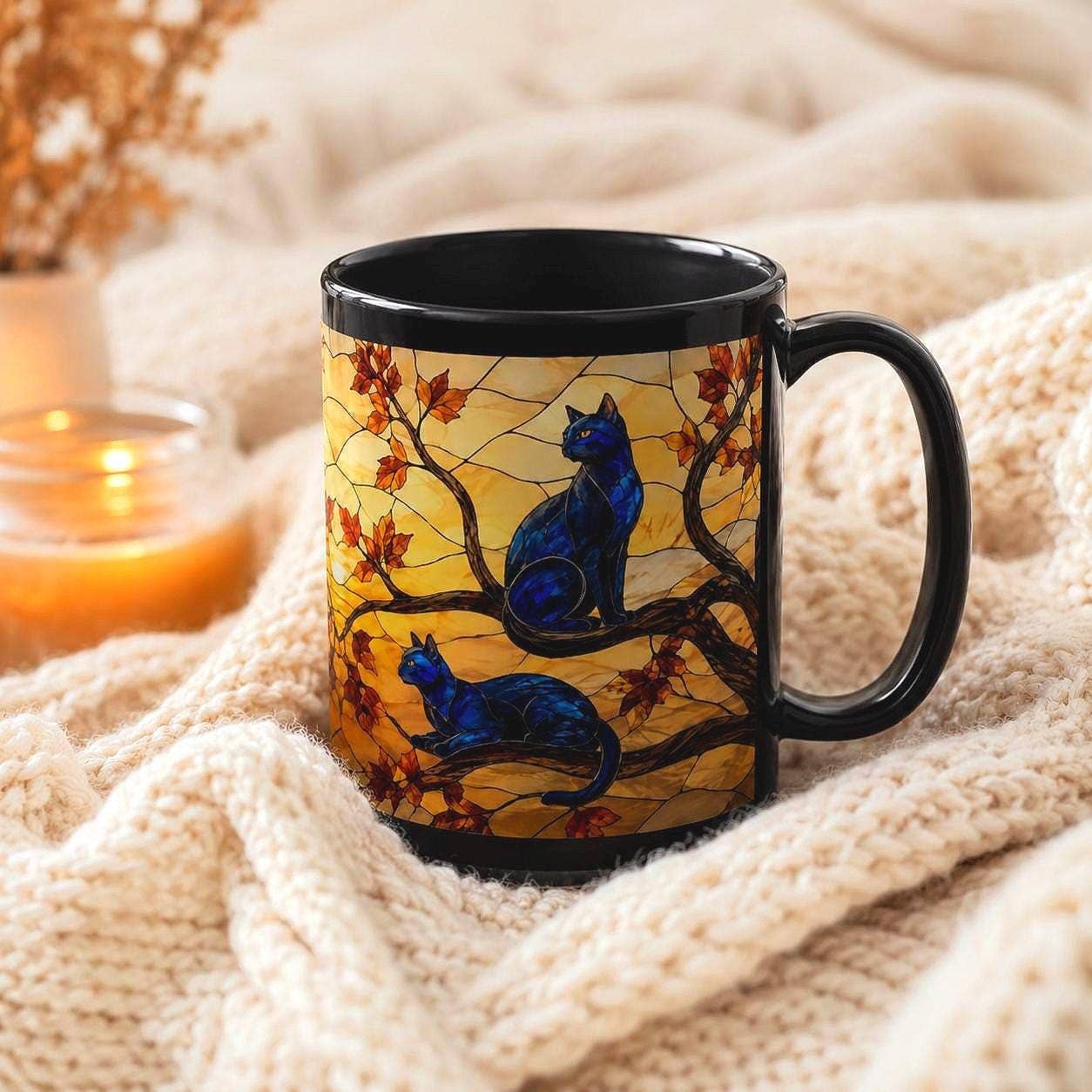A black ceramic mug with a printed design of two blue cats on a tree branch with autumn leaves, set on a cozy cream knit blanket with a warm candle in the background.