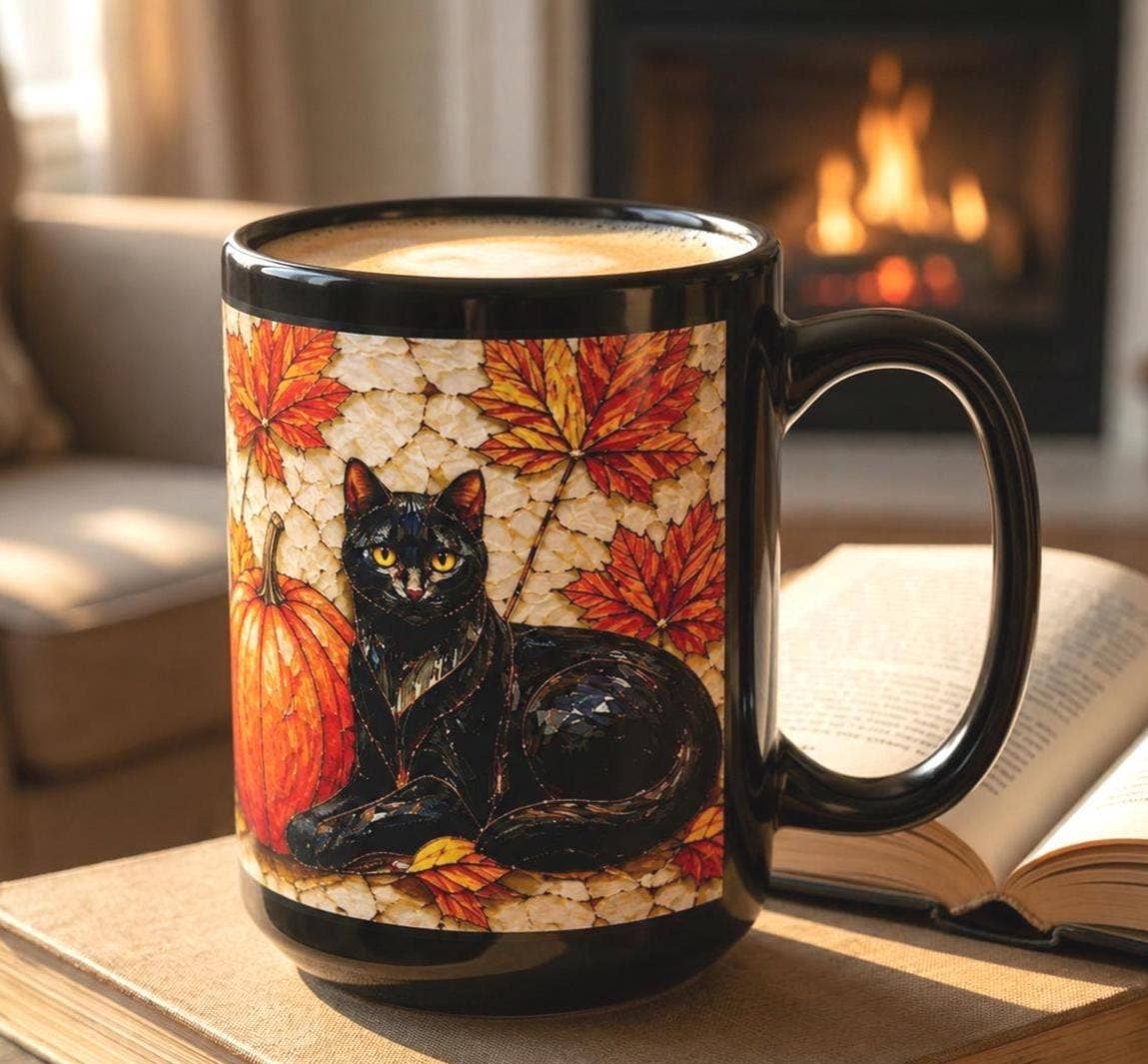 A black ceramic mug with a printed design of a black cat, pumpkins, and autumn leaves, sitting on a table in a cozy living room setting with a fireplace and an open book in the background.